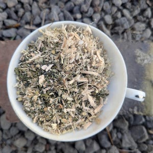 May include: A white teacup filled with a mix of dried herbs and plant matter. The contents are a blend of green, tan, and brown, with visible pieces of leaves and stems. The cup sits on a stone surface, with a background of small gray rocks.