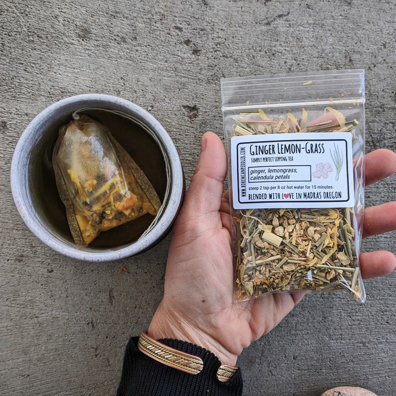 Puede incluir: Una bolsita de t&eacute; transparente llena de hierbas y flores secas, incluyendo jengibre, hierba lim&oacute;n y p&eacute;talos de cal&eacute;ndula. La bolsita de t&eacute; est&aacute; en una taza de cer&aacute;mica blanca. El t&eacute; est&aacute; etiquetado como "Ginger Lemon-Grass Simply Perfect Sipping Tea" e incluye instrucciones para preparar 2 cucharaditas por 237 ml de agua caliente durante 15 minutos. El t&eacute; se mezcla con amor en Madras, Oreg&oacute;n.