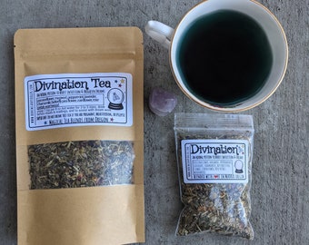 May include: An overhead shot features two pouches of "Divination Tea" and a teacup filled with blue liquid. The tea pouches are brown and clear, displaying dried herbs. The teacup is white with a gold rim, and a small purple crystal sits nearby.