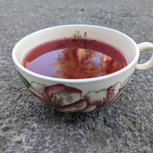 May include: A white ceramic teacup filled with a deep red liquid, possibly tea or a similar beverage. The cup features floral designs in shades of pink and green. The cup is resting on a gray, textured surface.