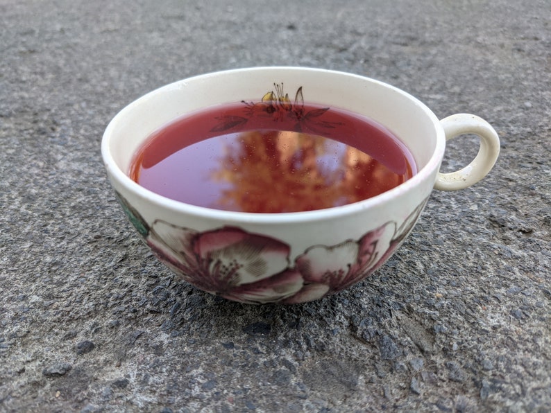 May include: A white ceramic teacup filled with a deep red liquid, possibly tea or a similar beverage. The cup features floral designs in shades of pink and green. The cup is resting on a gray, textured surface.