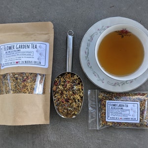 May include: A brown paper bag with a clear window showing a blend of dried flowers and herbs labeled "Flower Garden Tea" with the text "Blended with Love in Madras, Oregon". A metal scoop filled with the tea blend is next to the bag. A small clear bag with the same tea blend is on the right. A white teacup with a floral design is filled with light brown tea and is on a white saucer with a floral design.