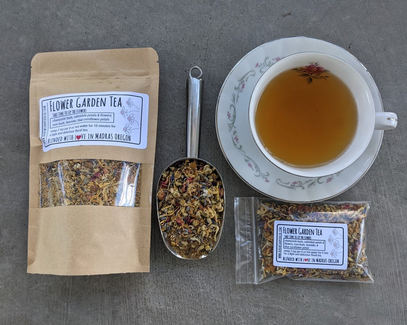 May include: A brown paper bag with a clear window showing a blend of dried flowers and herbs labeled "Flower Garden Tea" with the text "Blended with Love in Madras, Oregon". A metal scoop filled with the tea blend is next to the bag. A small clear bag with the same tea blend is on the right. A white teacup with a floral design is filled with light brown tea and is on a white saucer with a floral design.