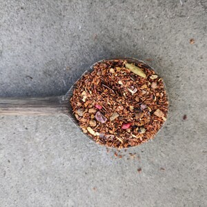 Op de afbeelding: Een houten lepel gevuld met een mengsel van losse bladthee. De thee is een mix van rode, bruine en gele stukjes, met wat kleine rode bessen en stukjes chocolade.