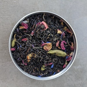 May include: A close-up of a metal tin filled with black tea leaves, dried rose petals, and other floral ingredients.