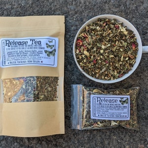 May include: Two brown paper bags with clear plastic windows filled with a blend of dried herbs and flowers. The bags are labelled "Release Tea" and include a list of ingredients and instructions. The text on the bags reads: "An herbal potion that brings emotions to the surface so you can let go and move forward." The bags are sitting on a grey, textured surface.