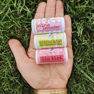 May include: Three small tubes of lip balm with floral infused lip balm labels. The labels are pink, green, and white with black text. The labels read "bohemian peddler's citrus lavender floral infused lip balm", "bohemian peddler's citrus mint floral infused lip balm", and "bohemian peddler's lip tint floral infused lip balm".