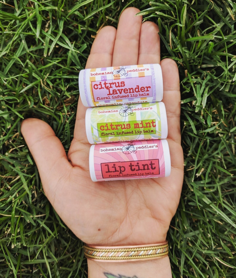 May include: Three small tubes of lip balm with floral infused lip balm labels. The labels are pink, green, and white with black text. The labels read "bohemian peddler's citrus lavender floral infused lip balm", "bohemian peddler's citrus mint floral infused lip balm", and "bohemian peddler's lip tint floral infused lip balm".