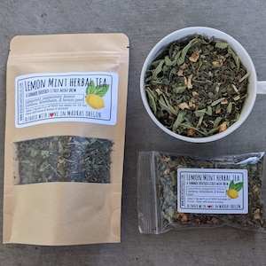May include: Two brown paper bags filled with loose leaf tea, one with a label that reads "Lemon Mint Herbal Tea A Summer Inspired Citrus Mint Brew Spearmint, Peppermint, Lemon Verbena, Lemon Balm, & Lemon Peel Steep 1 tsp per 8 oz of VERY hot water for 10 mins. Flavor with honey & lemon if desired. Blended with LOVE in Madras Oregon", and the other with a similar label. A white ceramic cup filled with the same tea is in the background.