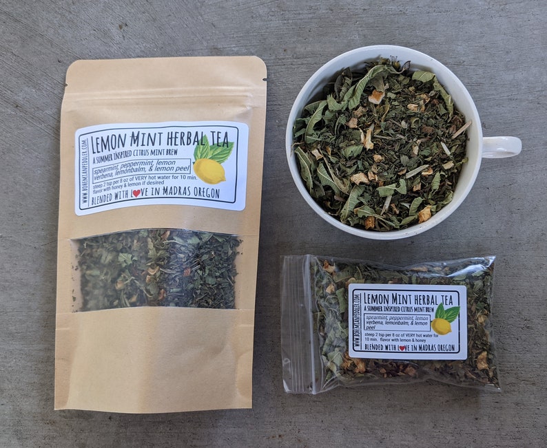 May include: Two brown paper bags filled with loose leaf tea, one with a label that reads "Lemon Mint Herbal Tea A Summer Inspired Citrus Mint Brew Spearmint, Peppermint, Lemon Verbena, Lemon Balm, & Lemon Peel Steep 1 tsp per 8 oz of VERY hot water for 10 mins. Flavor with honey & lemon if desired. Blended with LOVE in Madras Oregon", and the other with a similar label. A white ceramic cup filled with the same tea is in the background.