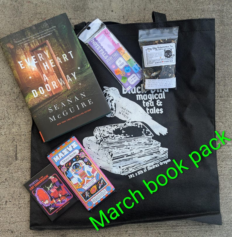 May include: A black tote bag containing books, tea, and stickers. The bag has a graphic of a bird and books, with text reading "Blackbird magical tea & tales." Books include "Every Heart a Doorway" and "Moon Rocks."