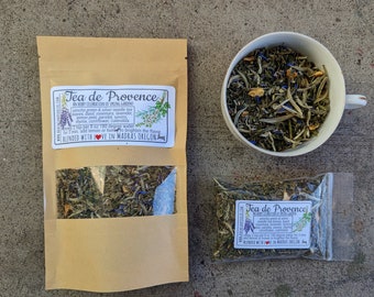 May include: A brown paper bag with a clear window showing a blend of green tea and herbs. The bag is labeled "Tea de Provence" and describes the ingredients as an "herbal celebration of spring gardens".
