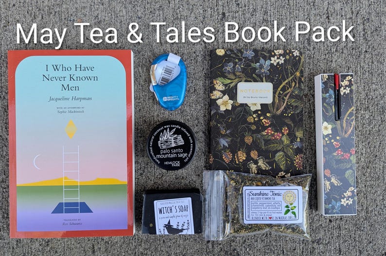 May include: A selection of items including a book titled "I Who Have Never Known Men", a floral notebook, a blue tape dispenser, a candle, a bar of soap, a bag of tea, and a pen set. The text "May Tea & Tales Book Pack" is at the top.