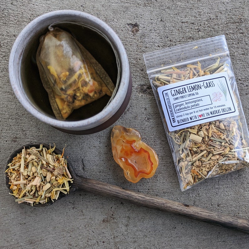 Puede incluir: Un primer plano de una taza de cer&aacute;mica marr&oacute;n con una bolsita de t&eacute; dentro. La bolsita de t&eacute; est&aacute; llena de hierbas y flores secas. La taza est&aacute; sobre una superficie gris. Hay una peque&ntilde;a bolsa de t&eacute; suelto al lado de la taza. La bolsa est&aacute; etiquetada como "Ginger Lemon-Grass" y contiene jengibre seco, hierba de lim&oacute;n y p&eacute;talos de cal&eacute;ndula. El texto en la bolsa dice "Simply Perfect Supporting Tea" y "Infuse 2 cucharaditas por 8 oz de agua caliente durante 15 minutos. Mezclado con amor en Madras Oregon."