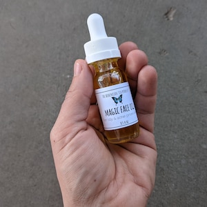 May include: A small glass bottle with a white dropper top and a white label. The label has a blue butterfly graphic and the text "The Bohemian Tea Producer Magic Face Oil for oily & acne prone skin 0.5 fl oz".