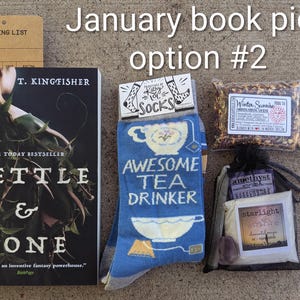 May include: A gift set including a book by T. Kingfisher, socks with "Awesome Tea Drinker" text, tea, and a bag with "starlight" text. The set is labelled "January book pick option #2". The book is titled "Nettle & Bone".