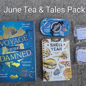 May include: A flat lay of a "June Tea & Tales Pack" featuring a book titled "Voyage of the Damned", a sardine tin, a sticker book, and two tea packets. The book is blue with gold lettering. The sticker book is white with colorful shell illustrations.
