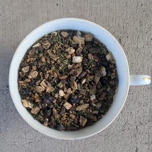 May include: A white ceramic bowl filled with a blend of dried herbs and berries for tea. The blend includes dried ginger, juniper berries, and other herbs.