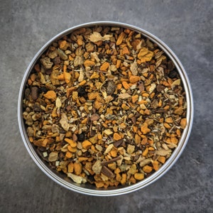 May include: A close-up of a metal tin filled with a blend of dried herbs and spices, including turmeric, ginger, and cinnamon. The mixture is a vibrant orange color and appears to be a tea blend.