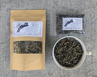 May include: A brown paper bag with a clear window showing a blend of dried herbs and flowers. The bag has a label that reads "The Crone's Cooling Cup" and lists the ingredients: red clover, raspberry leaves, peppermint, chamomile, lady's mantle, lemon balm, alfalfa, ginger, sage, passionflower, hibiscus, and calendula. The label also states "Blended with Love in Madras, Oregon".