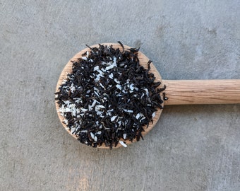 May include: A wooden spoon filled with black tea leaves and white coconut flakes. The spoon is positioned diagonally against a gray concrete surface. The tea and coconut are the primary focus, showcasing their texture and color contrast.