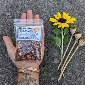 May include: A clear plastic bag of September Spice tea blend held in a hand, with a yellow sunflower and dried seed pods. The bag has a label with text and a tree illustration. The tea blend includes hibiscus, orange peel, clove, cinnamon, star anise, and safflower.