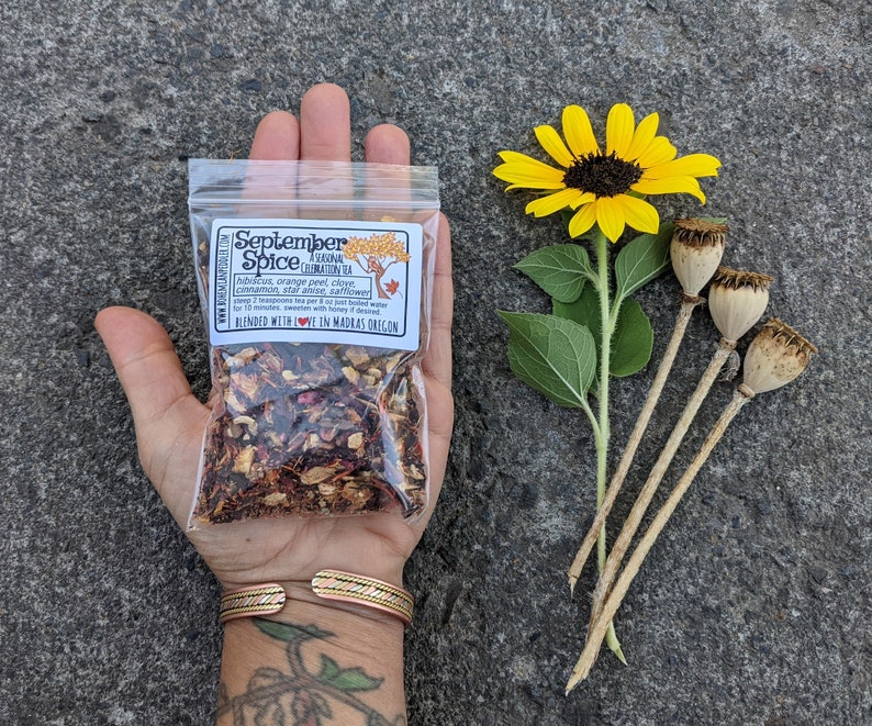 May include: A clear plastic bag of September Spice tea blend held in a hand, with a yellow sunflower and dried seed pods. The bag has a label with text and a tree illustration. The tea blend includes hibiscus, orange peel, clove, cinnamon, star anise, and safflower.