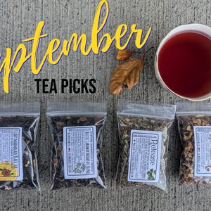 May include: Four clear plastic pouches containing loose-leaf tea blends labeled Lady Marmalade, Rustic Caraway, Release, and September Spice, alongside a white teacup filled with dark red tea and two dried brown leaves, with the text September Tea Picks.