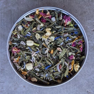 May include: A close-up of a metal container filled with a blend of green, yellow, and purple dried herbs and flowers. The mixture includes green tea leaves, lavender, rose petals, and other botanicals.