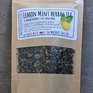 May include: A brown paper bag with a clear window showing a blend of dried herbs for a lemon mint herbal tea. The bag has a label that reads "Lemon Mint Herbal Tea" and lists the ingredients: spearmint, peppermint, lemon, verbena, lemon balm, and lemon peel. The label also says "A Summer Inspired Citrus Mint Brew" and "Blended with Love in Madras Oregon".