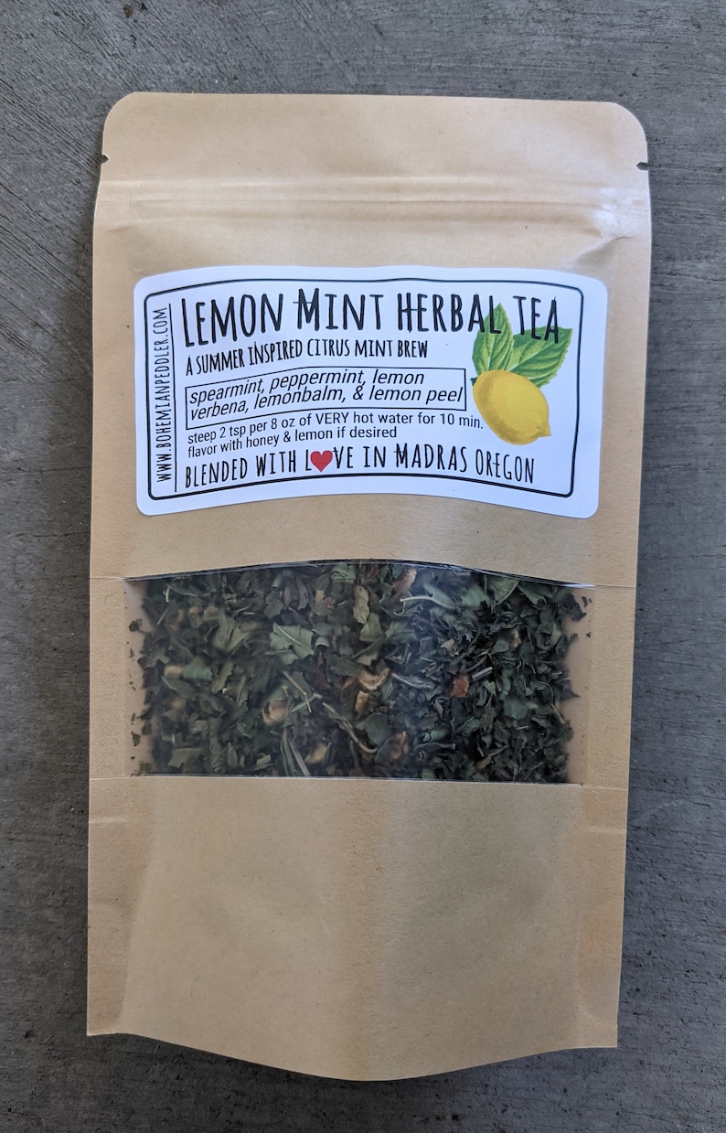 May include: A brown paper bag with a clear window showing a blend of dried herbs for a lemon mint herbal tea. The bag has a label that reads "Lemon Mint Herbal Tea" and lists the ingredients: spearmint, peppermint, lemon, verbena, lemon balm, and lemon peel. The label also says "A Summer Inspired Citrus Mint Brew" and "Blended with Love in Madras Oregon".