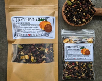 May include: Two brown paper bags with a label that reads "Orange Chocolate Chai" and a description of the ingredients. The bags contain a blend of black tea, orange peel, cocoa nibs, ginger, allspice, cinnamon, cardamom, and black pepper. The bags are sitting on a wooden surface.