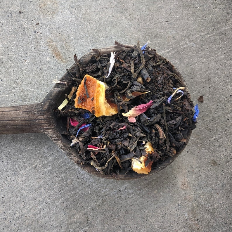 Puede incluir: Una cuchara de madera llena de mezcla de t&eacute; negro de hoja suelta. El t&eacute; incluye c&aacute;scara de naranja seca, p&eacute;talos de flores azules y rosas y hojas de t&eacute; negro.