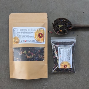 Puede incluir: Una bolsa de papel marr&oacute;n con una ventana transparente que muestra t&eacute; negro con flores secas, c&aacute;scara de naranja y especias. La bolsa tiene una etiqueta que dice "Lady Marmalade Black Tea" con una descripci&oacute;n del t&eacute; y las palabras "Blended with Love in Madras Oregon".
