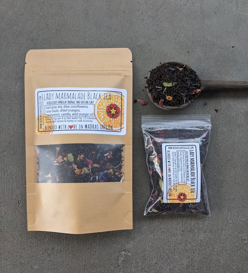 Puede incluir: Una bolsa de papel marr&oacute;n con una ventana transparente que muestra t&eacute; negro con flores secas, c&aacute;scara de naranja y especias. La bolsa tiene una etiqueta que dice "Lady Marmalade Black Tea" con una descripci&oacute;n del t&eacute; y las palabras "Blended with Love in Madras Oregon".