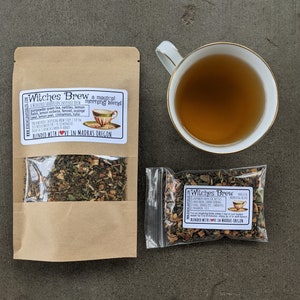 May include: Two brown paper bags with clear plastic windows containing loose leaf tea blends. The bags are labeled "Witches Brew a magical morning blend" and list the ingredients. The tea blends are green, brown, and orange in color.