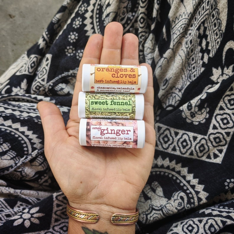 May include: Three small lip balm tubes stacked on top of each other. The tubes are labelled "oranges & cloves", "sweet fennel", and "candy ginger". Each tube has a floral infused lip balm description.