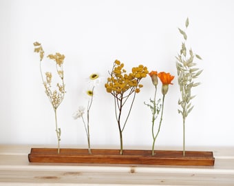 Könnte beinhalten: Eine Holz-Blumenvase mit sechs Schlitzen für getrocknete Blumen. Die Vase ist mit getrockneten Blumen in den Farben Weiß, Gelb und Orange gefüllt.