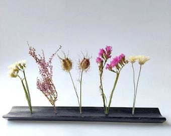 Può includere: Un vaso di fiori in legno nero con cinque fessure per gli steli. Il vaso è pieno di fiori secchi, tra cui fiori bianchi, rosa e marroni.