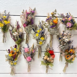 Può includere: Una collezione di dieci bouquet di fiori secchi, ciascuno legato con spago. I bouquet presentano una varietà di fiori ed erbe colorate, tra cui fiori gialli, viola, rosa e arancioni. I bouquet sono disposti su una superficie di legno bianco.