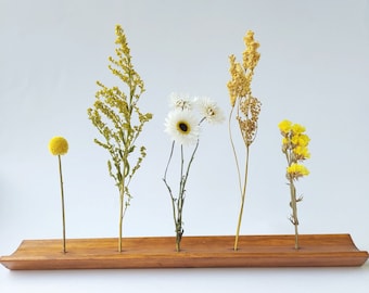 Può includere: Un vaso di fiori in legno con cinque fessure per gli steli. Il vaso è pieno di fiori secchi in tonalità di giallo e bianco. I fiori includono piccoli pompon gialli, margherite bianche e rametti gialli.