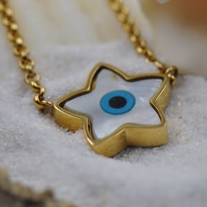 Puede incluir: Collar dorado con un colgante en forma de estrella con incrustaciones de nácar y un diseño de ojo azul. El collar está sobre un fondo blanco y arenoso, resaltando los detalles de la joya. La cadena también es dorada.