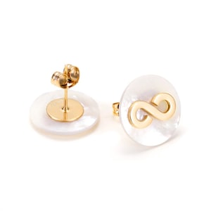 Puede incluir: Un par de pendientes de botón dorados. Cada pendiente presenta una base circular blanca de nácar con un símbolo de infinito dorado. Los postes y cierres de los pendientes también son dorados. Los pendientes están sobre un fondo blanco.