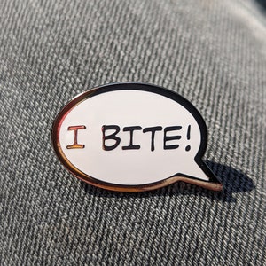 May include: A pin in the shape of a speech bubble with the words "I BITE!" in black and red lettering. The pin has a white background, a black outline, and a gold-colored border. The pin is resting on a gray denim surface.