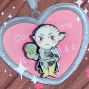 May include: A silver enamel pin featuring a cartoon character with pointed ears, a black and white outfit, and a green and purple orb in their hand.