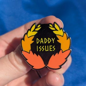 May include: A round enamel pin with the text "DADDY ISSUES" in yellow lettering. The pin features a black background with a wreath design in yellow and orange. The pin is held in a hand.
