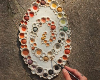 May include: A white ceramic artist's palette with a spiral design, filled with various colors of paint. The palette is held by a person with a tattoo, who is using a paintbrush to mix the colors. The palette is on a stone surface.