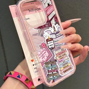 May include: A pink phone case with a clear camera lens cover, decorated with book-themed stickers and the Instagram logo. The stickers include phrases like "Dare Romance" and "Enemies to Lovers". A pink studded bracelet is visible in the foreground.