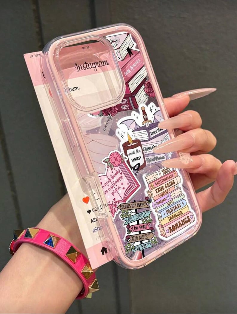 May include: A pink phone case with a clear camera lens cover, decorated with book-themed stickers and the Instagram logo. The stickers include phrases like "Dare Romance" and "Enemies to Lovers". A pink studded bracelet is visible in the foreground.