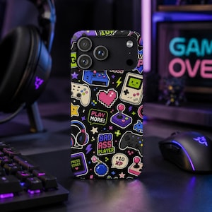 May include: Black phone case with a pixelated gaming theme. The case features colorful icons of game controllers, hearts, and text that says "PLAY MORE!" and "BAD ASS PLAYER". The case is on a desk with a keyboard, mouse, and headset.